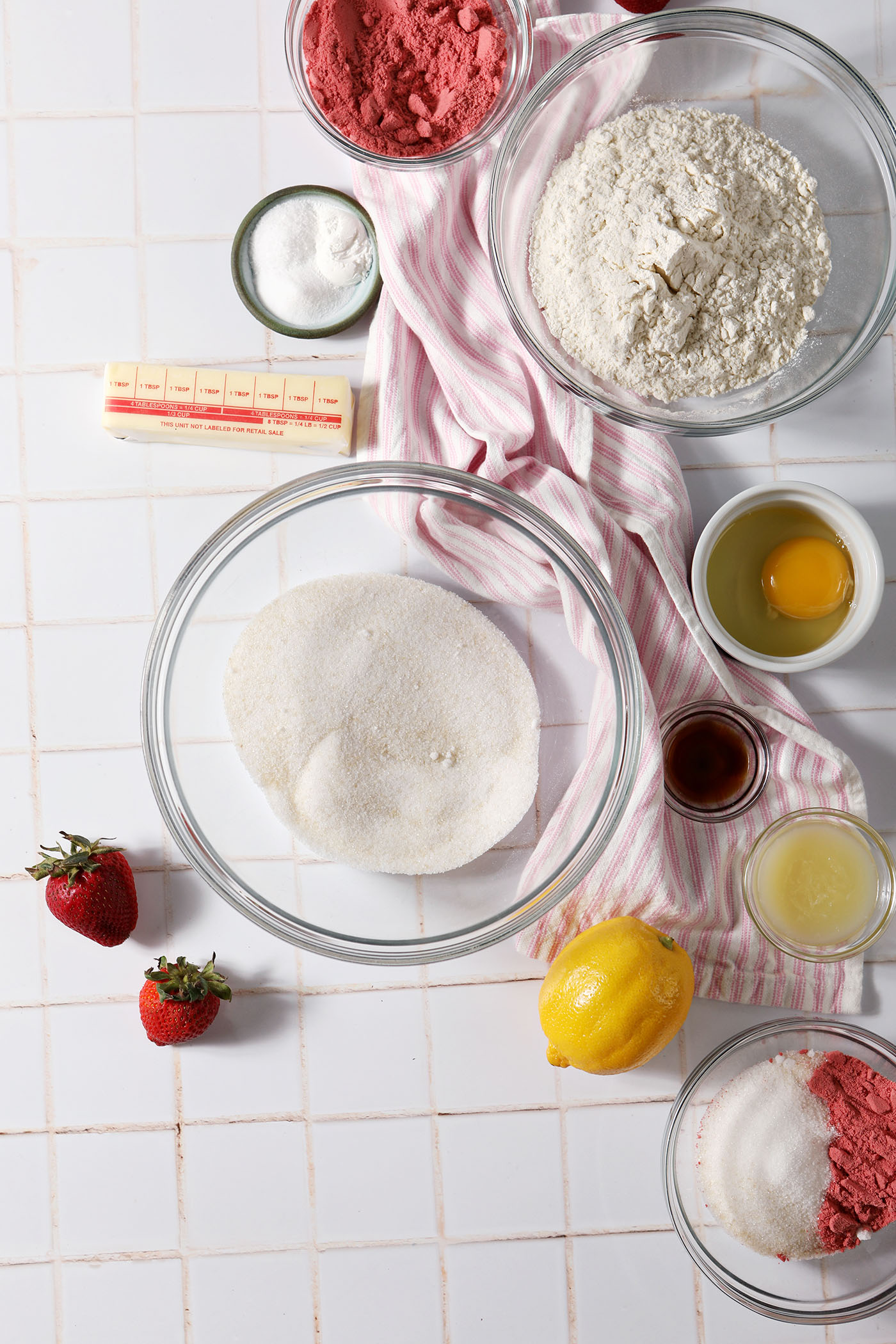 Ingredients to make lemon strawberry cookies in bowls on a white tiled surface next to a pink and white striped linen and fresh strawberries