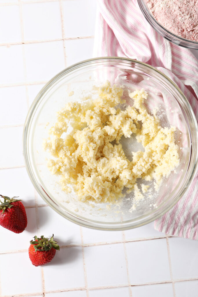 Creamed butter, sugar and lemon zest in a bowl on a white tiled surface next to strawberries and a linen