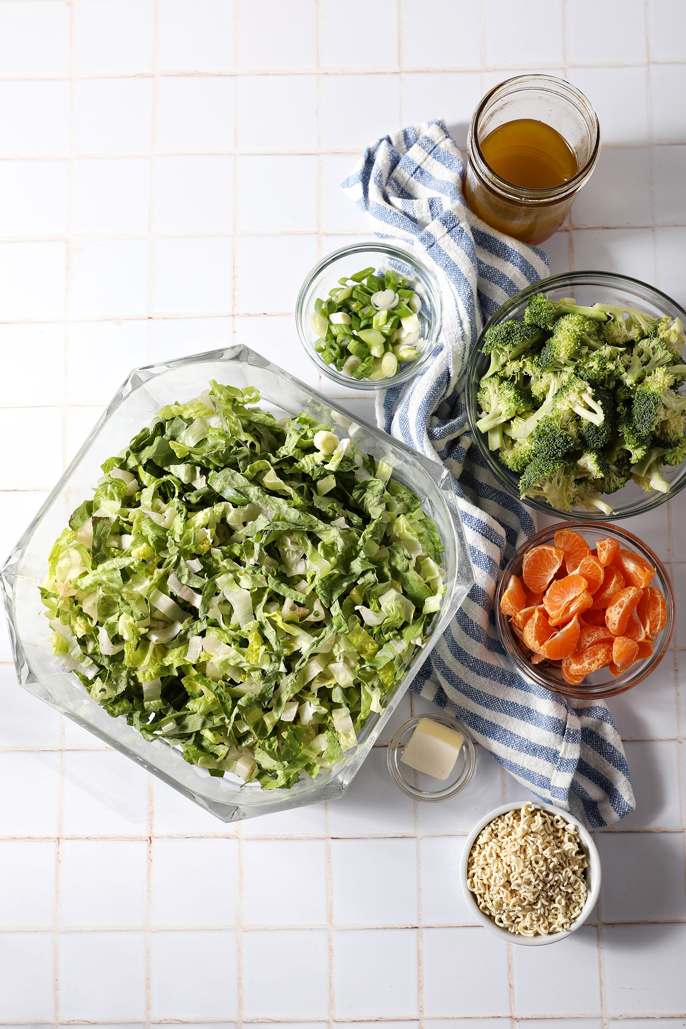 Ingredients to make a Ramen Noodle Salad with Clementines and Broccoli in bowls on a white tiled surface next to a blue and white striped linen