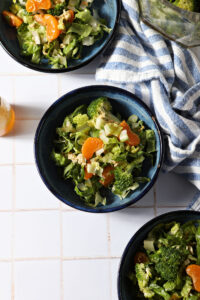 Three blue bowls holding clementine ramen noodle salad with broccoli on a white tiled surface next to a blue and white linen