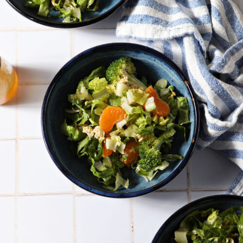 Three blue bowls holding clementine ramen noodle salad with broccoli on a white tiled surface next to a blue and white linen