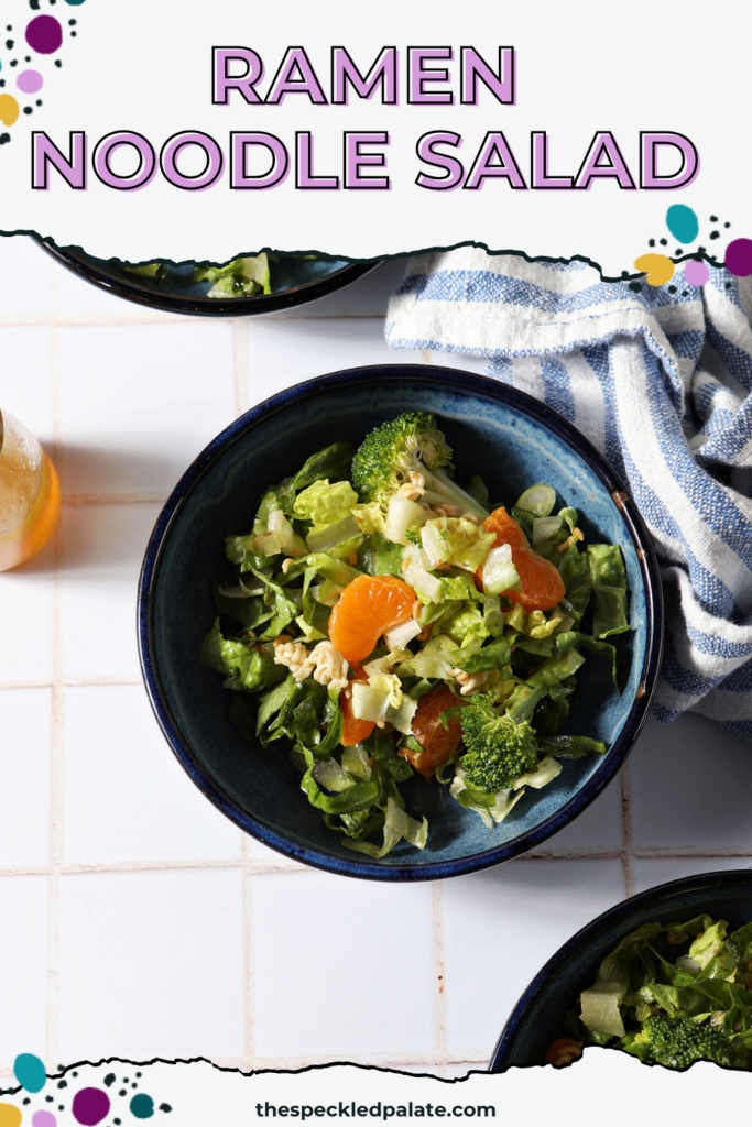 Close up of a blue bowl holding clementine ramen noodle salad with broccoli on a white tiled surface next to a blue and white linen with the text ramen noodle salad
