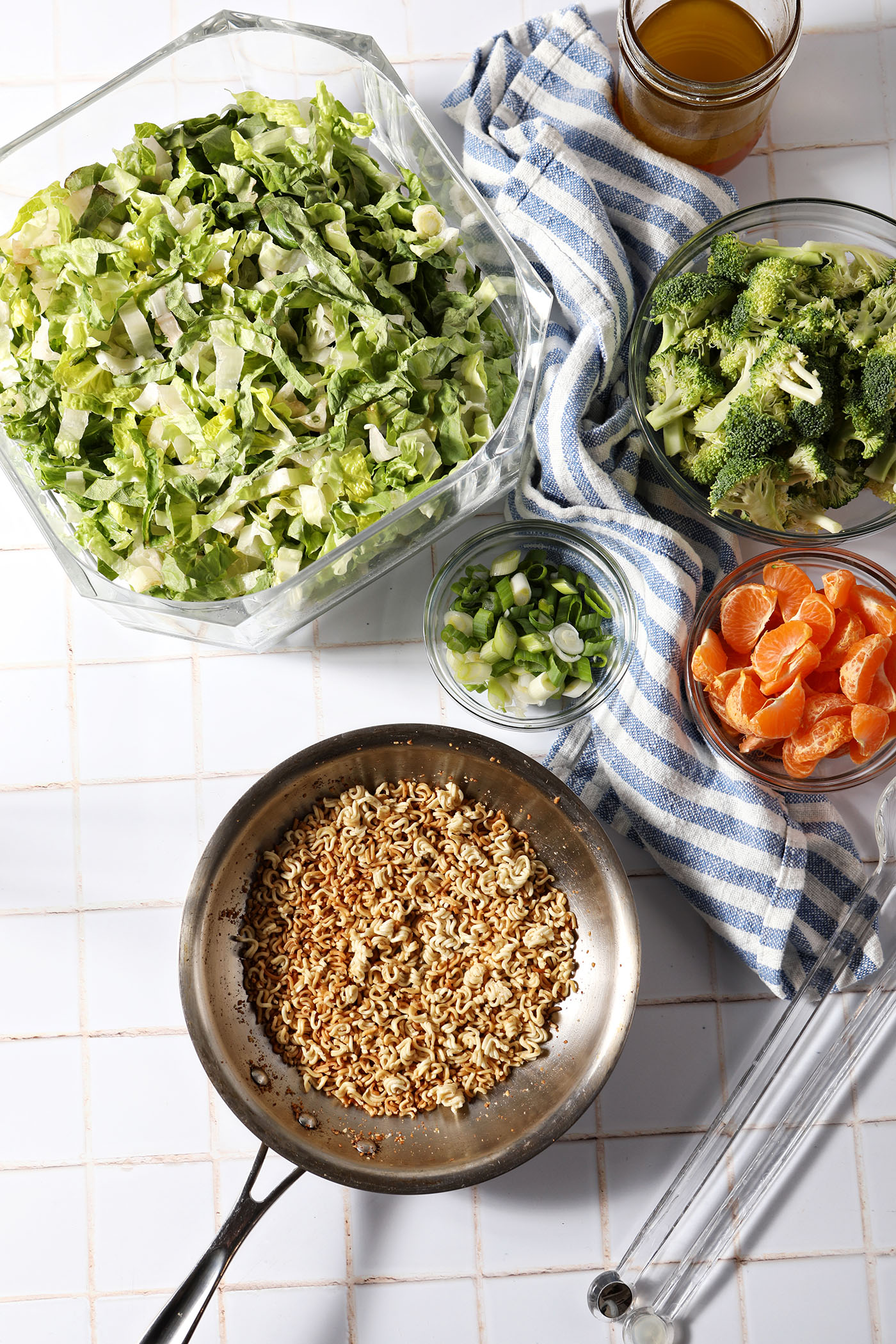 Ingredients to make Ramen Noodle Salad with Clementines and Broccoli in bowls and in a skillet on a white tiled surface before mixing together