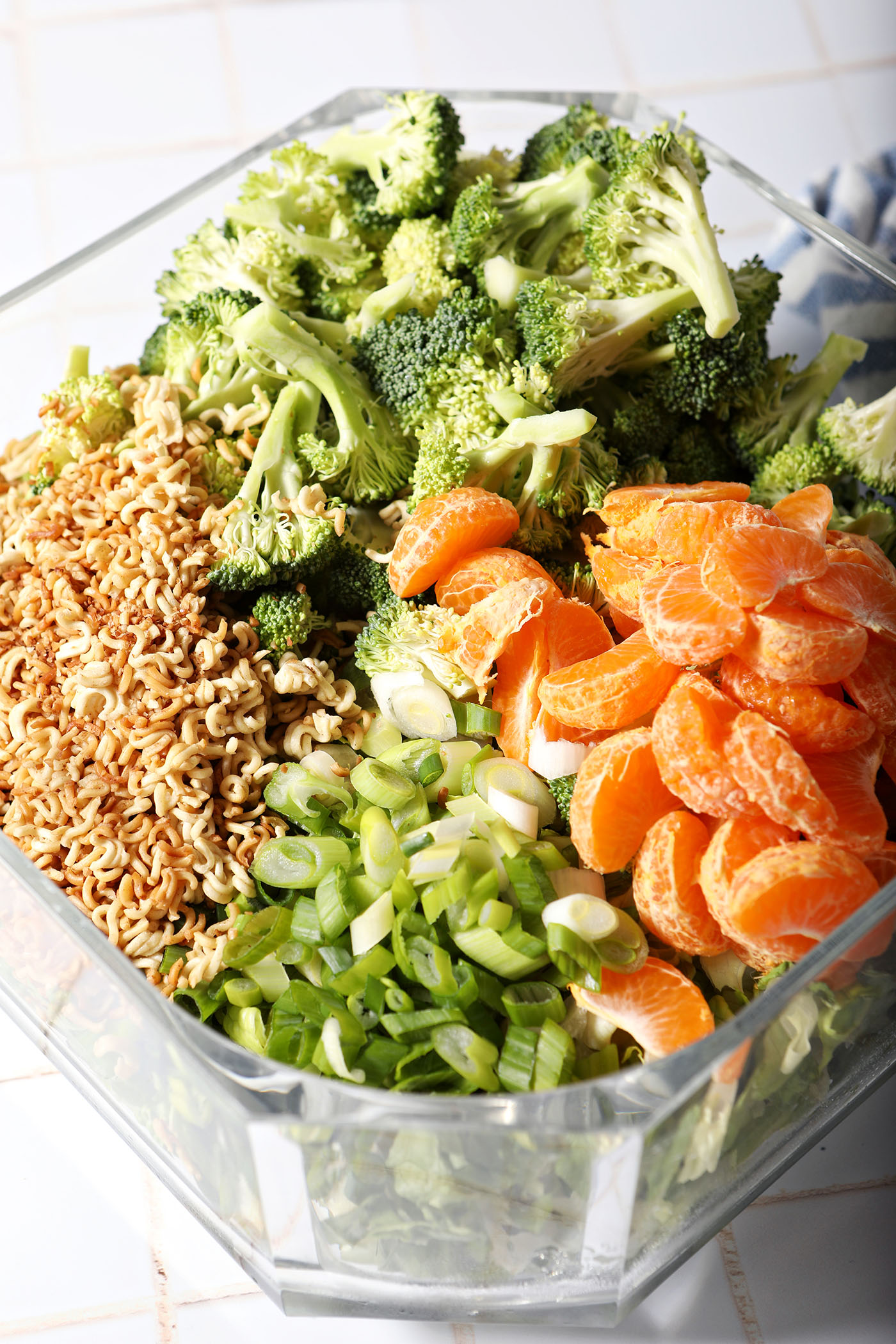 Toppings for a ramen noodle salad with clementines and broccoli sit on top of greens before tossing in a large salad bowl