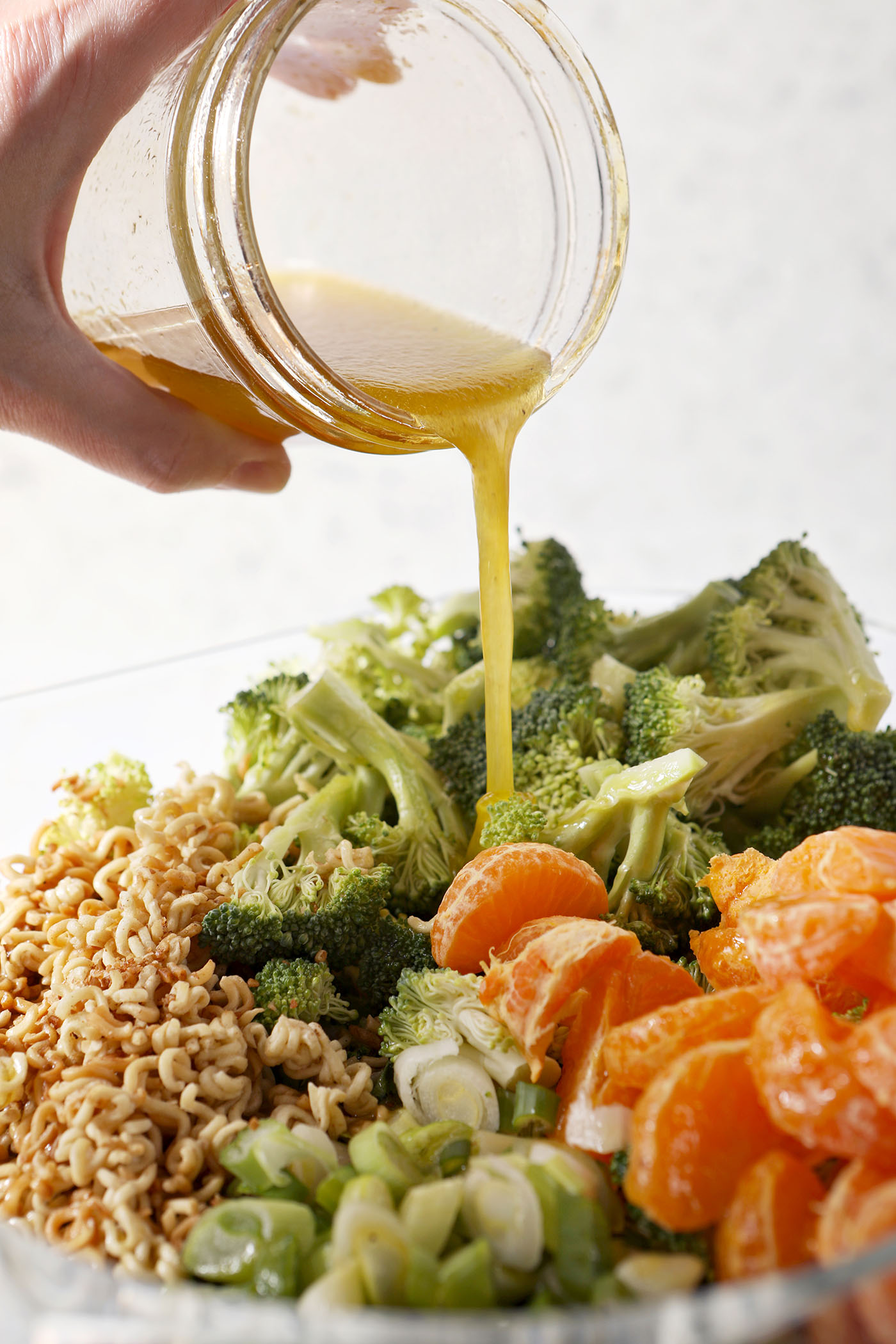 A mason jar of honey vinaigrette pours onto Ramen Noodle Salad with Clementines and Broccoli in a large salad bowl