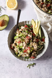 Overhead of a bowl holding a quinoa salad with chicken and veggies next to an avocado half and a lemon