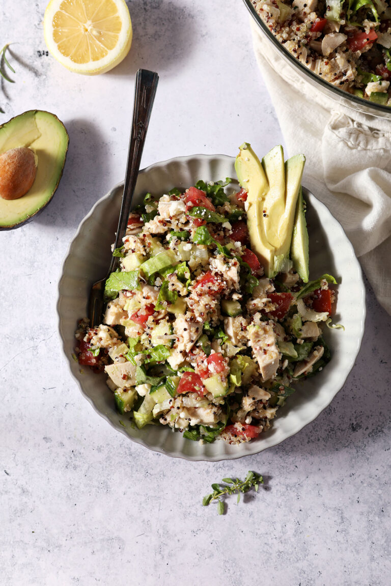 Overhead of a bowl holding a quinoa salad with chicken and veggies next to an avocado half and a lemon