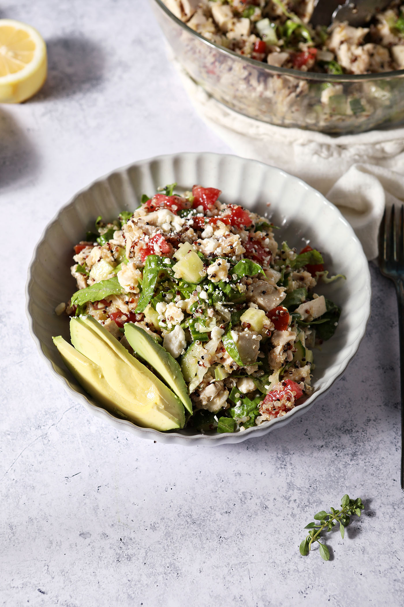 Side angle image of a bowl holding a lemon quinoa salad garnished with fresh herbs and sliced avocado in front of a larger salad bowl and a lemon