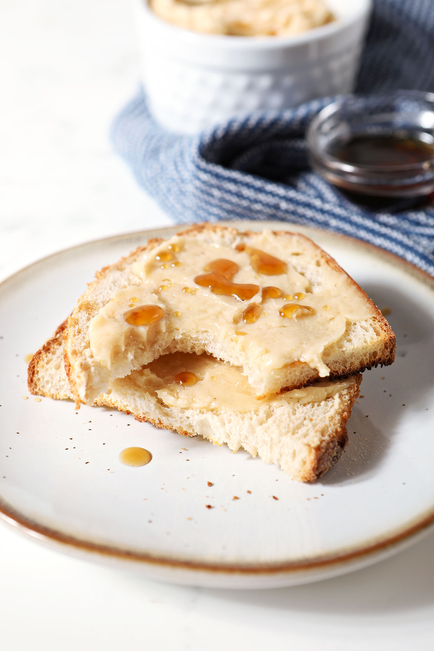 A halved slice of bread covered in maple butter on a plate in front of a blue linen and more butter and a container of maple syrup