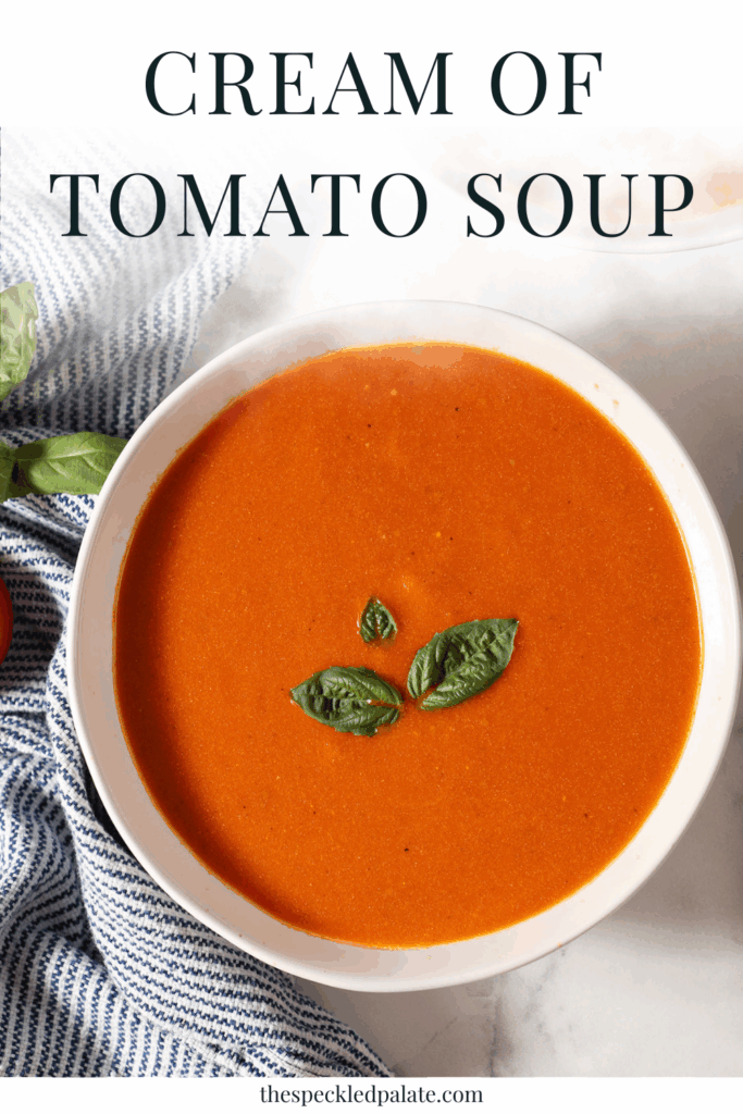 Close up of a bowl of tomato soup topped with fresh basil leaves on a marble surface next to a blue and white striped linen with the text cream of tomato soup