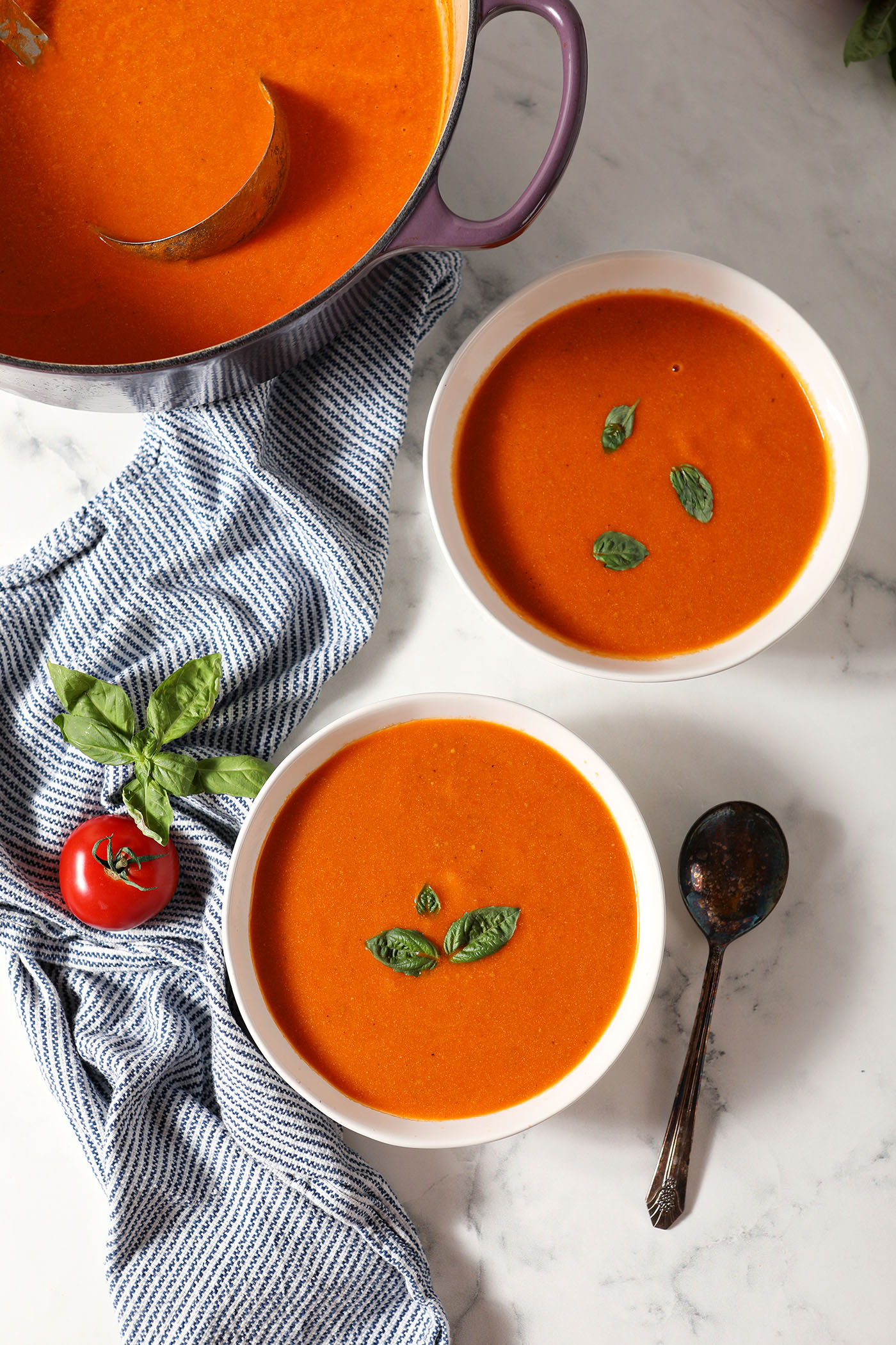 Two bowls of creamy tomato soup next to a dutch oven of soup with a blue and white linen and tomatoes and basil on a marble surface