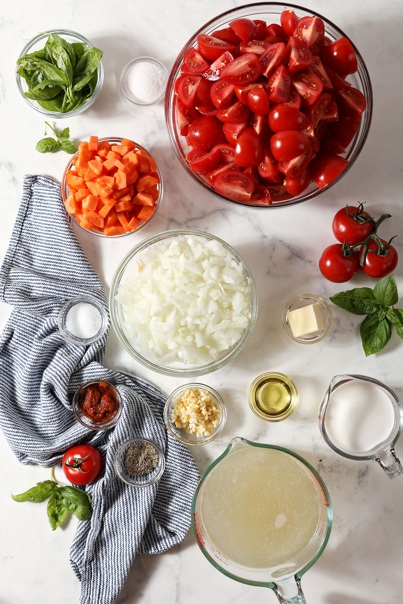Overhead image of ingredients to make fresh tomato soup with cream in bowls on a marble surface next to a blue and white linen
