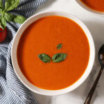 Close up of a bowl of fresh tomato soup with cream topped with fresh basil leaves on a marble surface next to a blue and white striped linen and another bowl
