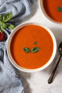 Close up of a bowl of fresh tomato soup with cream topped with fresh basil leaves on a marble surface next to a blue and white striped linen and another bowl