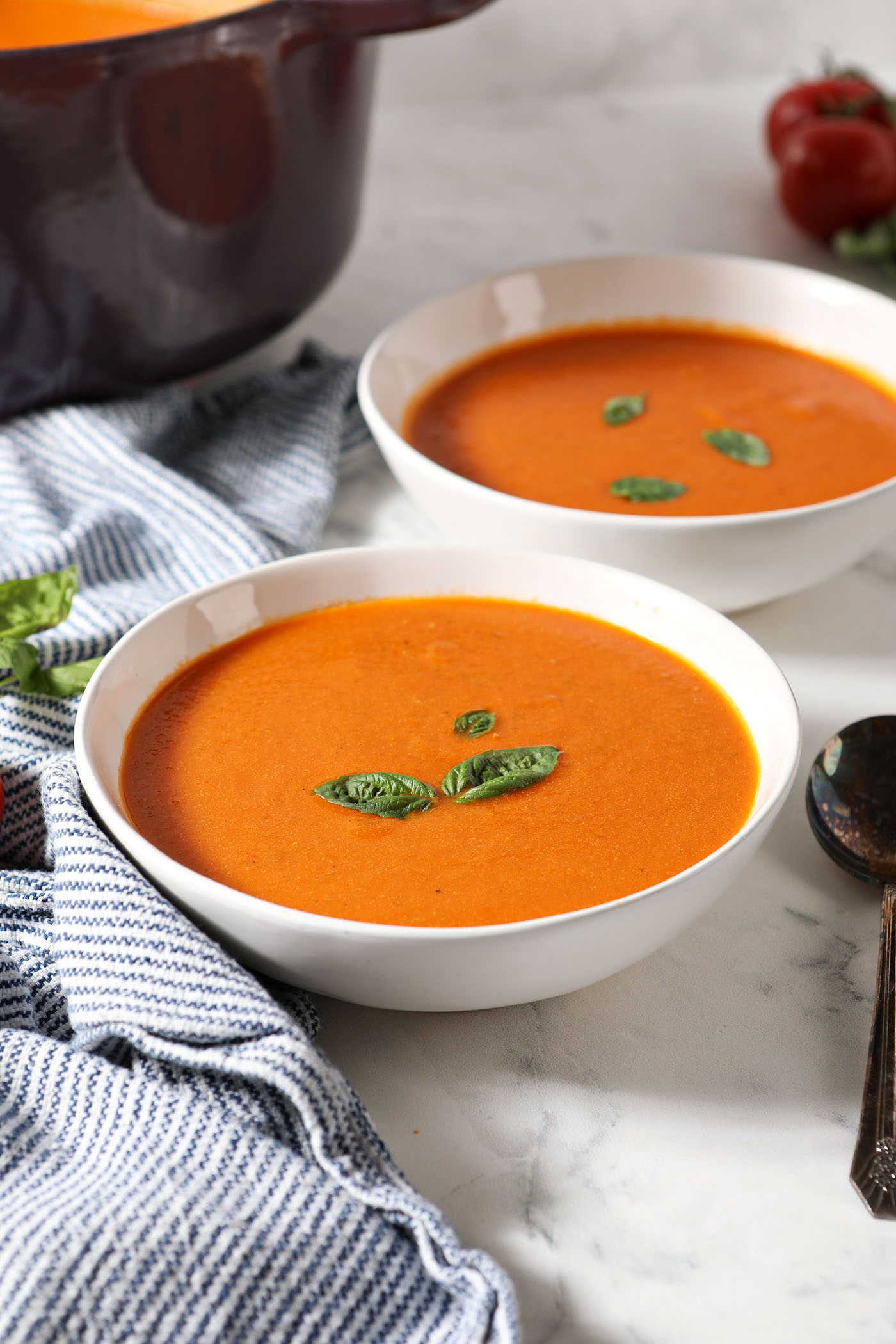 Side angle of two bowls of fresh cream of tomato soup next to a purple dutch oven, a blue and white striped linen and tomatoes and basil on a marble surface