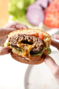Hands hold a juicy Lucy burger that is bursting with melted cheese
