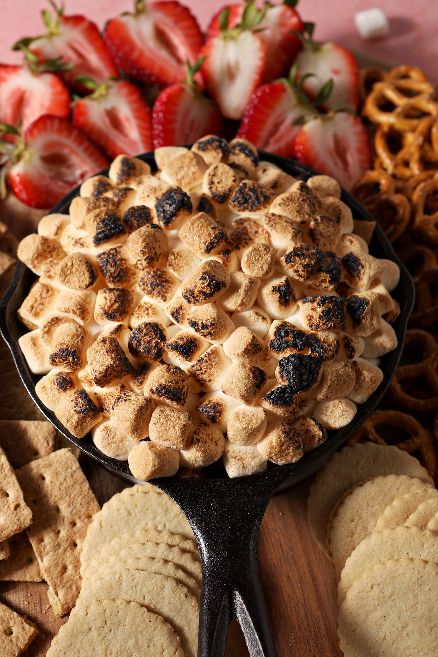 A skillet of baked smores dip surrounded by cookies, crackers and sliced strawberries