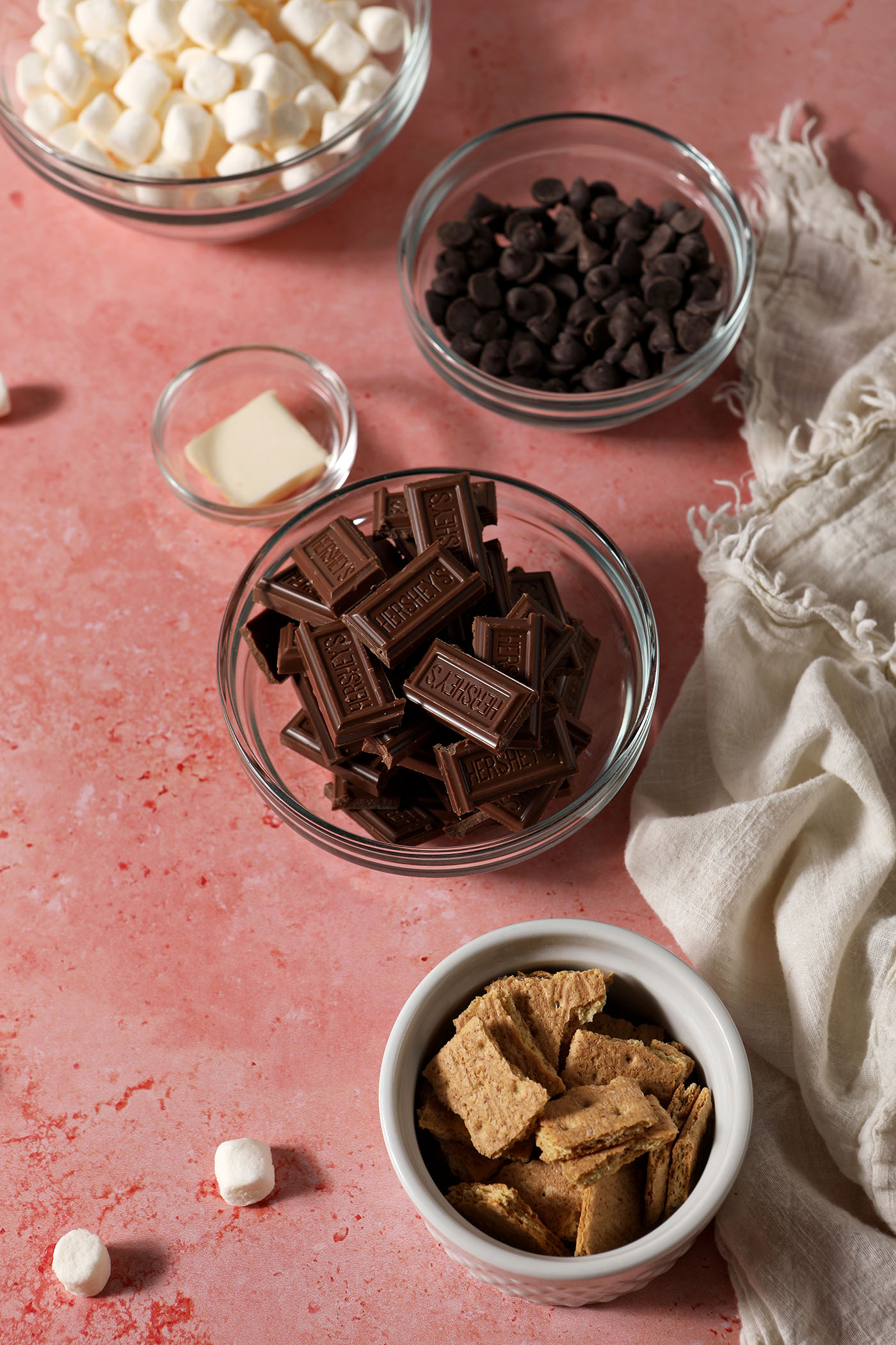 Ingredients to make s’mores dip in bowls on a pink surface with a white linen