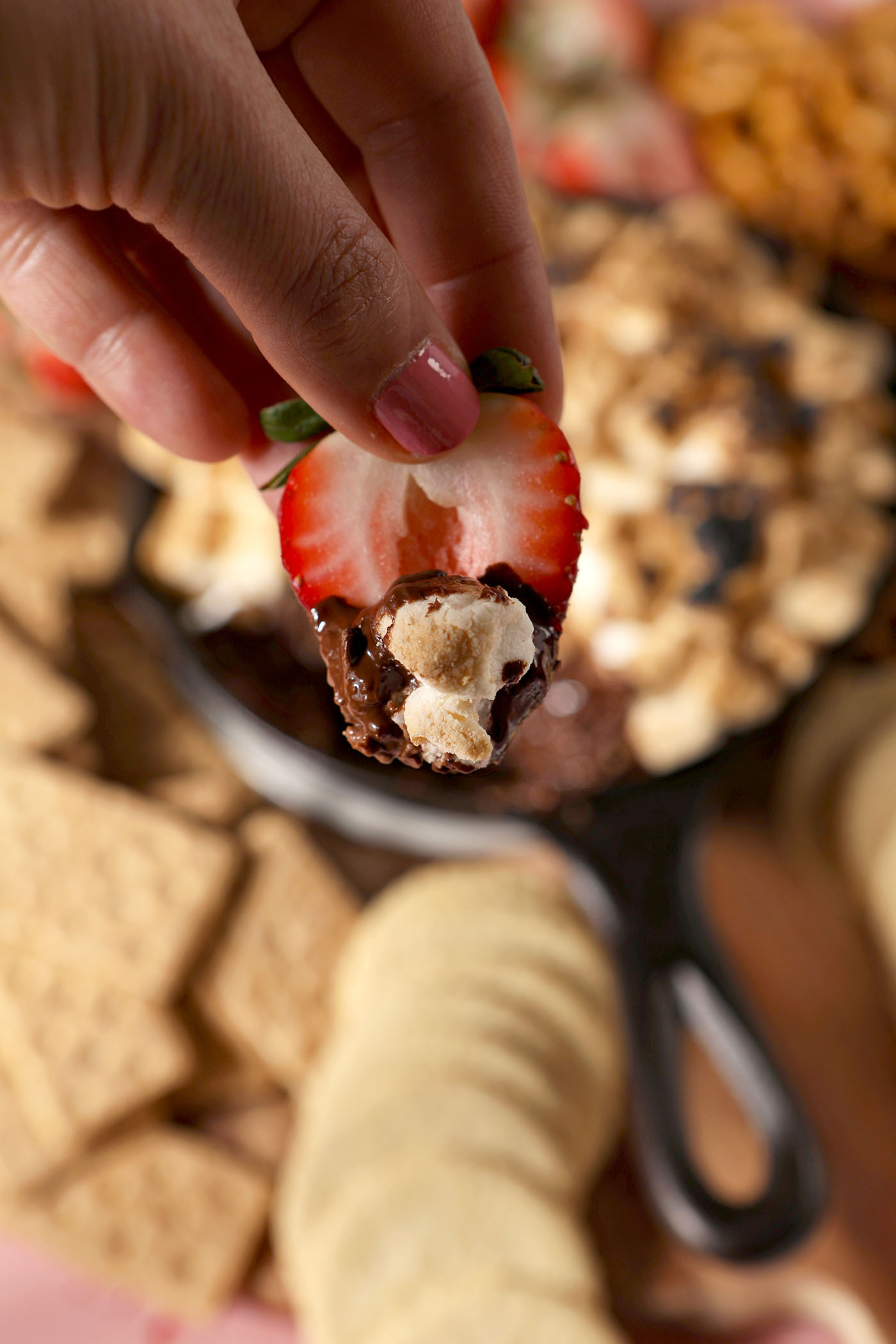 A hand holds a halved strawberry covered in s’mores dip above a board with a skillet of the dessert dip and more dippers