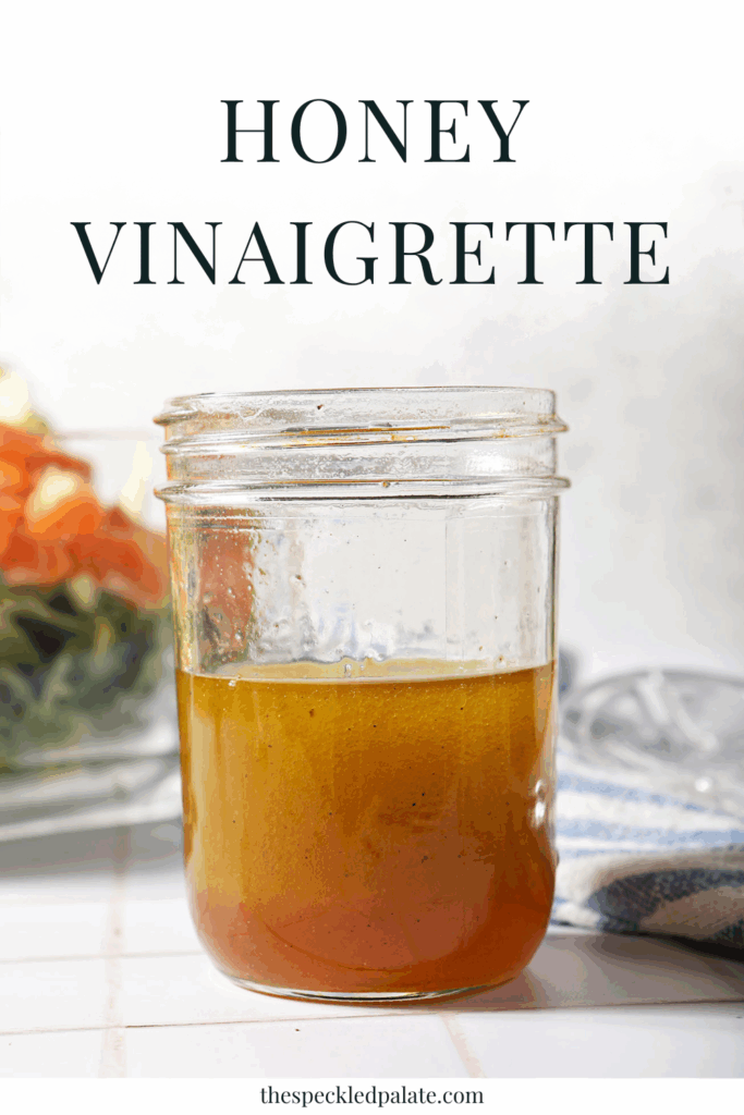 A jar of homemade honey salad dressing in front of a salad on a white tiled surface with the text honey vinaigrette