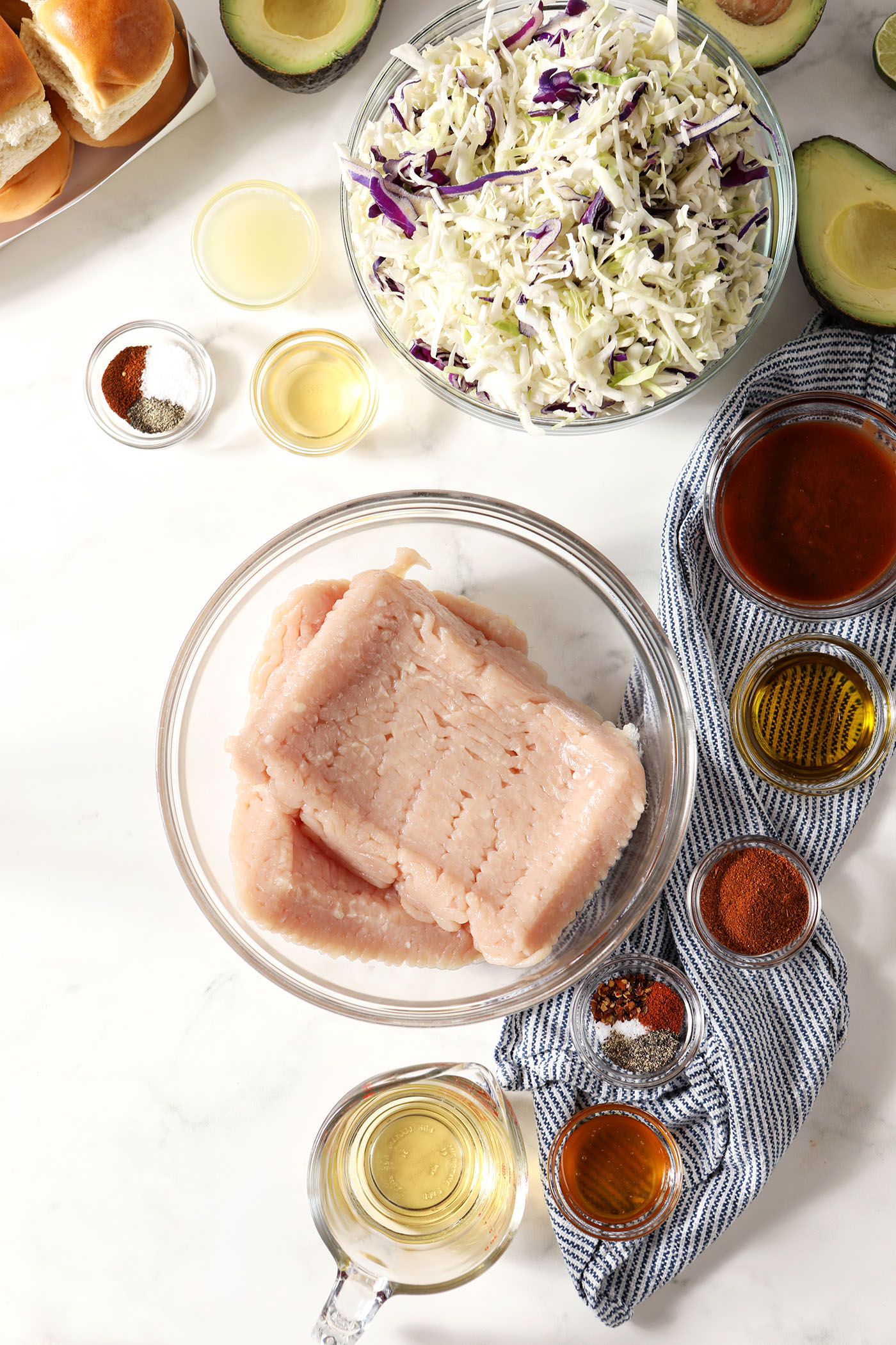Ingredients to make bbq chicken sliders and avocado coleslaw in bowls on a white surface