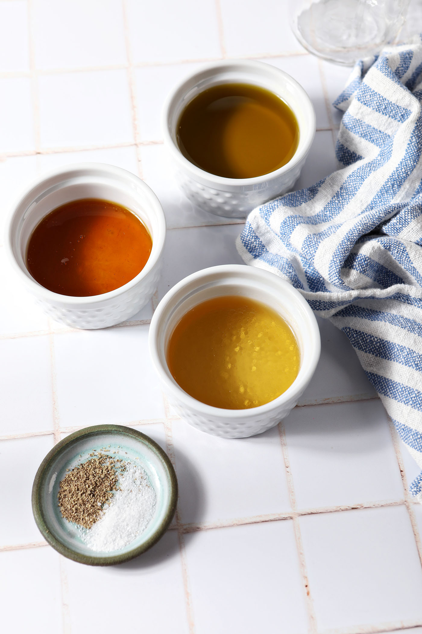 Ingredients to make a honey vinaigrette in bowls on a tiled surface next to a blue and white striped linen