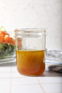 A jar of homemade honey vinaigrette in front of a salad on a white tiled surface
