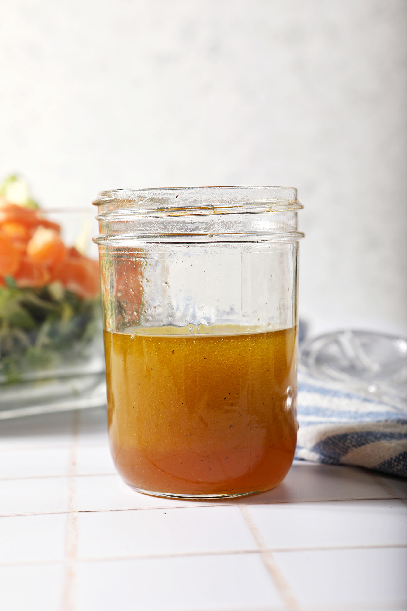 A jar of homemade honey vinaigrette in front of a salad on a white tiled surface