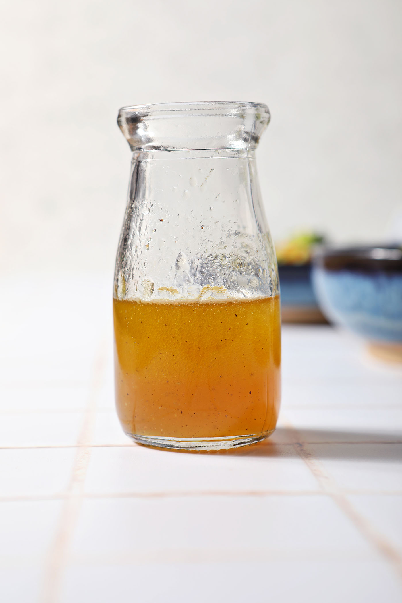 A bottle of homemade honey vinaigrette salad dressing on a white tiled surface in front of blue bowls
