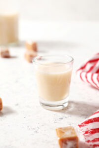 A shot glass holds a shot of caramel vodka on a textured white surface next to a white and red striped linen surrounded by soft caramels