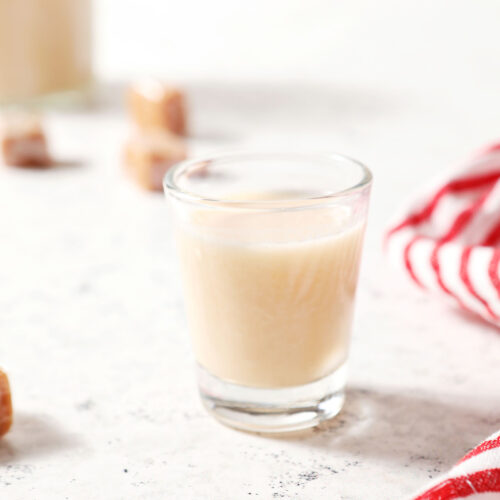 A shot glass holds a shot of caramel vodka on a textured white surface next to a white and red striped linen surrounded by soft caramels
