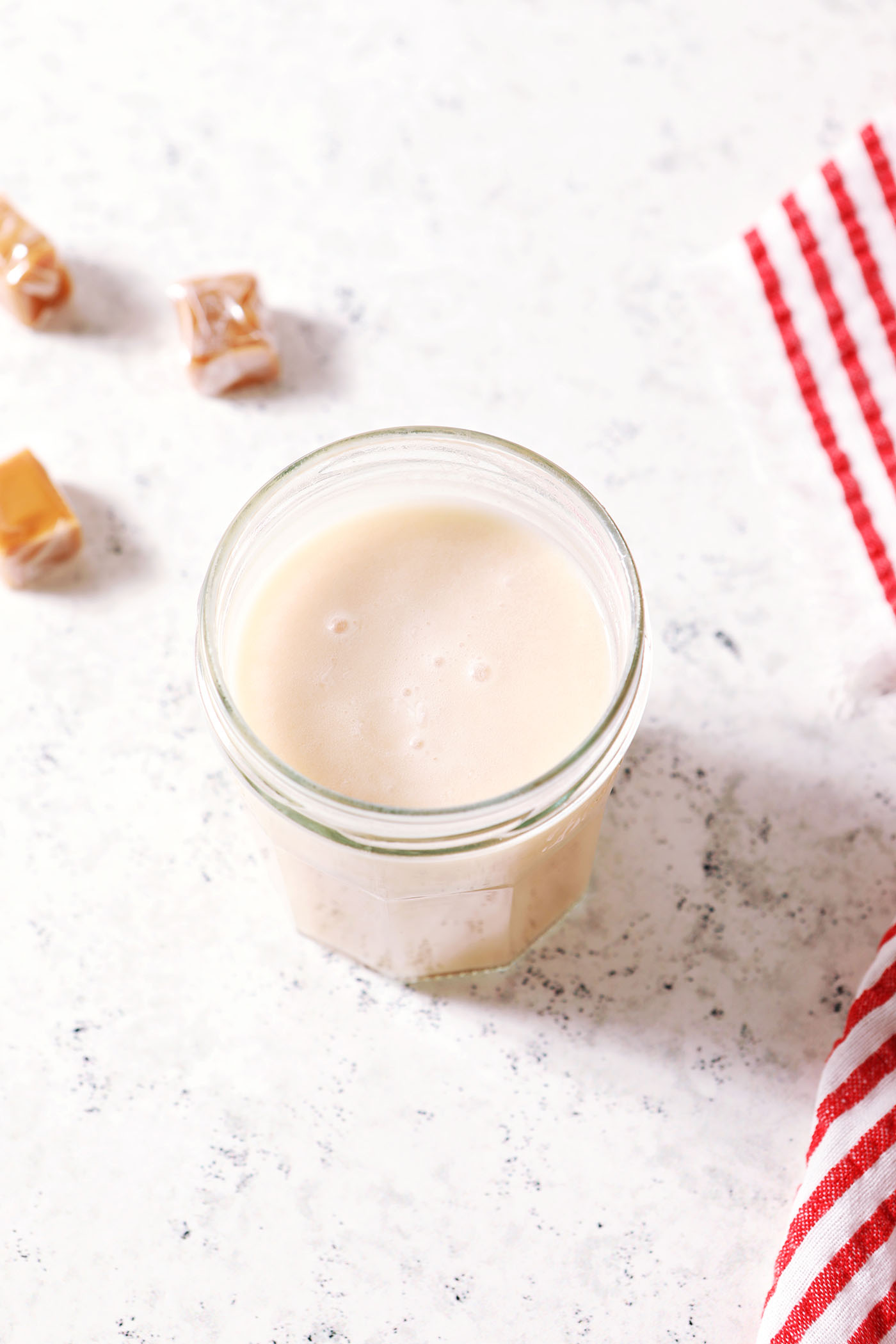 Overhead of a jar of homemade caramel infused vodka on a textured white surface next to a white and red striped linen surrounded by soft caramels