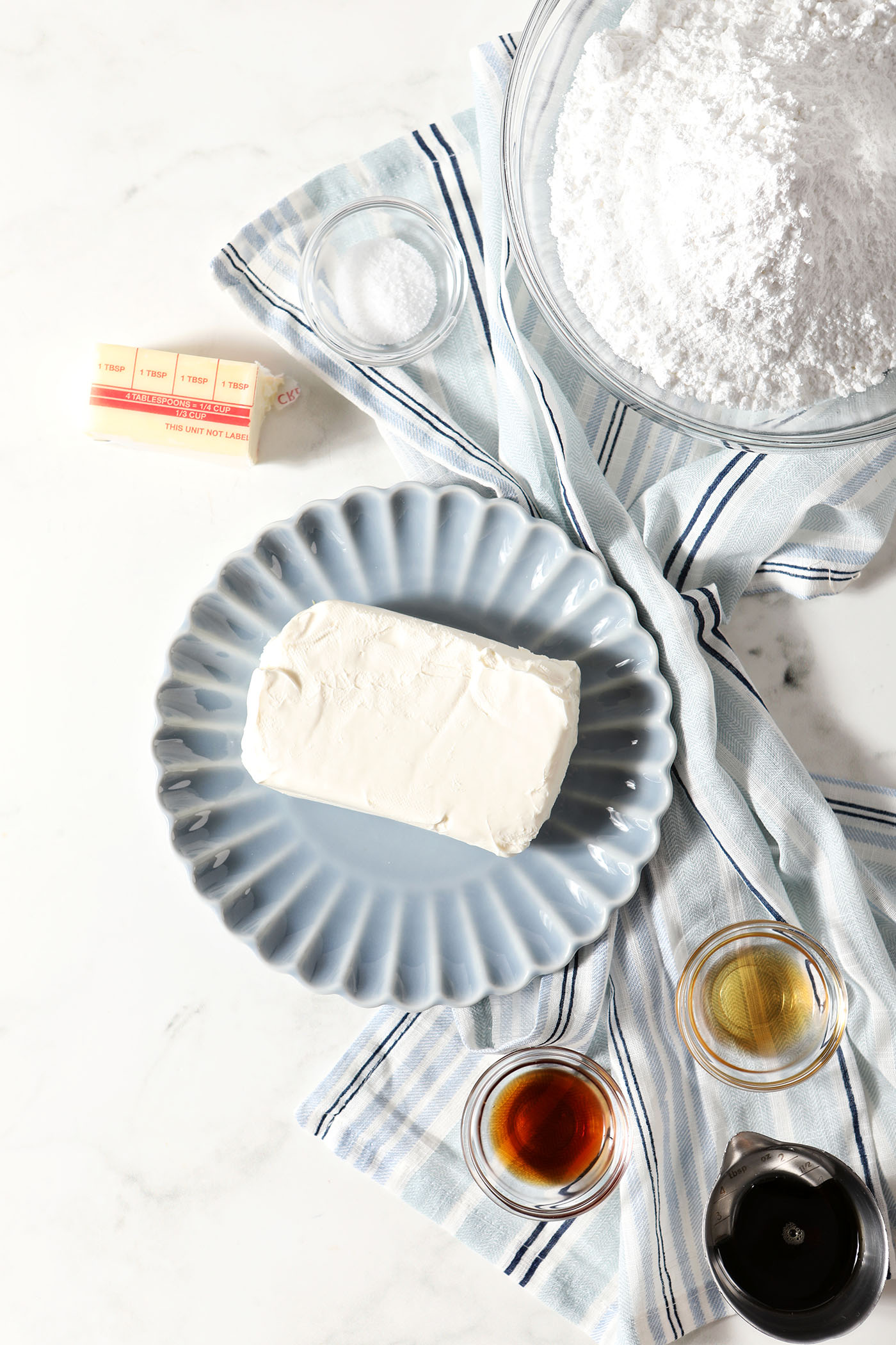 Ingredients to make cream cheese frosting with maple in bowls and plates on a stone surface next to a blue and white striped linen