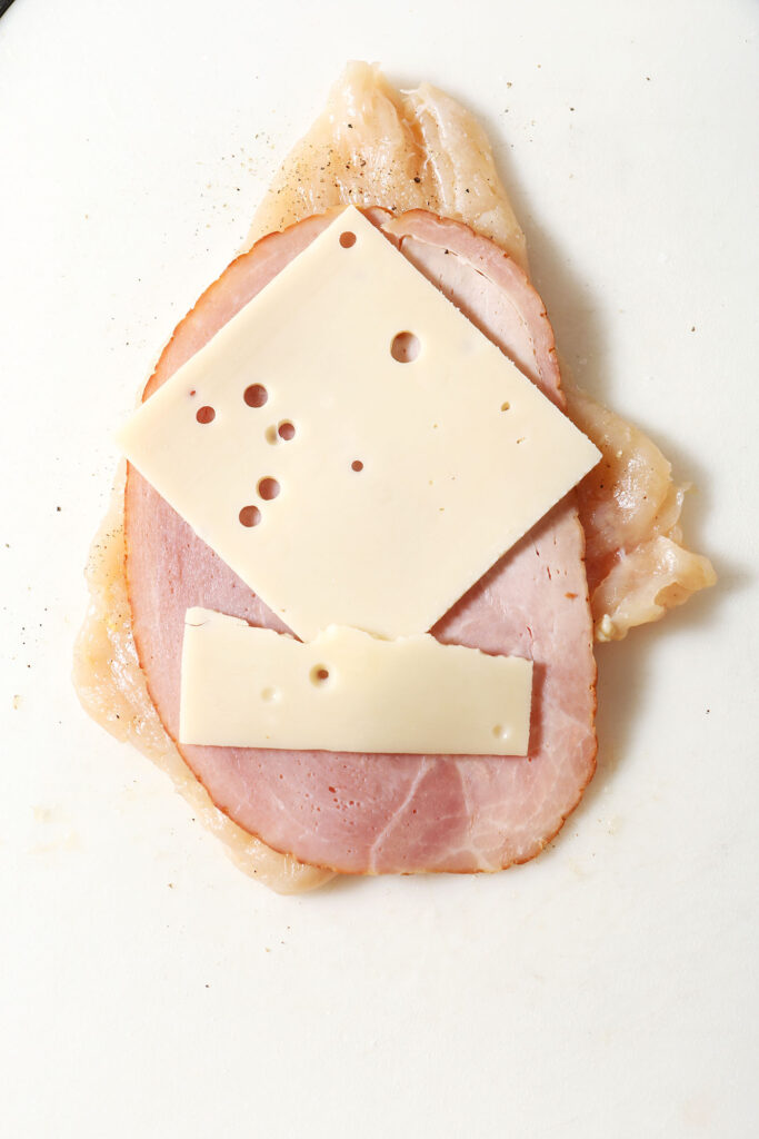 A slices of cheese and ham on top of a tenderized chicken breast