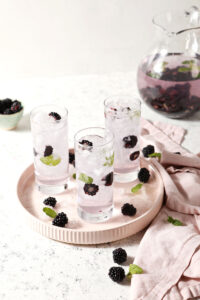 A pink platter holds three glasses of spa water with blackberries and mint in front of a pitcher of the slaw water on a stone surface next to pink linens