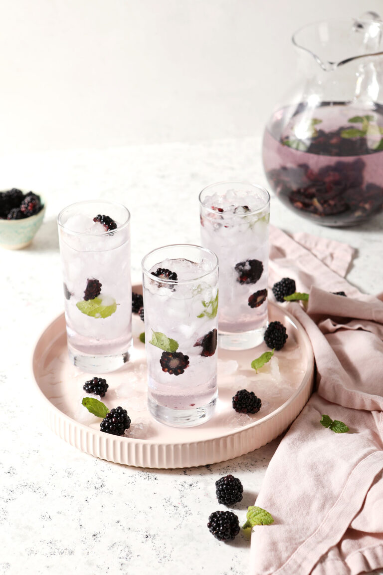 A pink platter holds three glasses of spa water with blackberries and mint in front of a pitcher of the slaw water on a stone surface next to pink linens