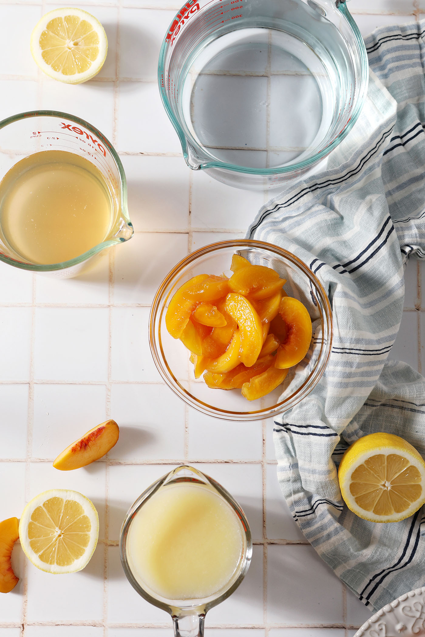 Ingredients to make homemade peach lemonade from above on a white tiled surface with a blue striped linen