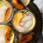 Overhead of three glasses of peach lemonade in a metal bowl with ice on a white tiled surface with a blue striped linen