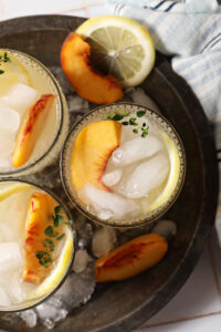 Overhead of three glasses of peach lemonade in a metal bowl with ice on a white tiled surface with a blue striped linen