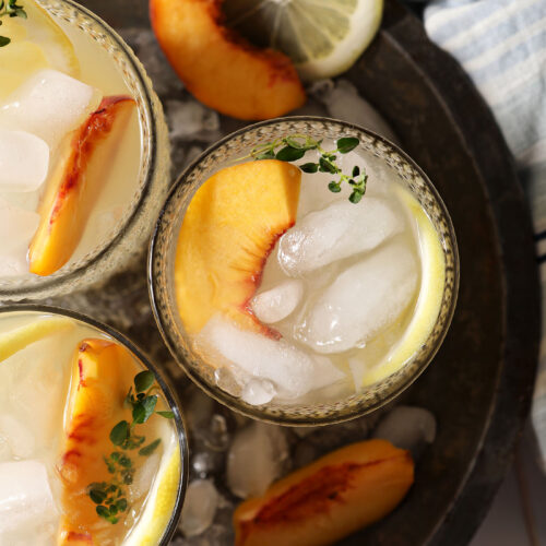 Overhead of three glasses of peach lemonade in a metal bowl with ice on a white tiled surface with a blue striped linen