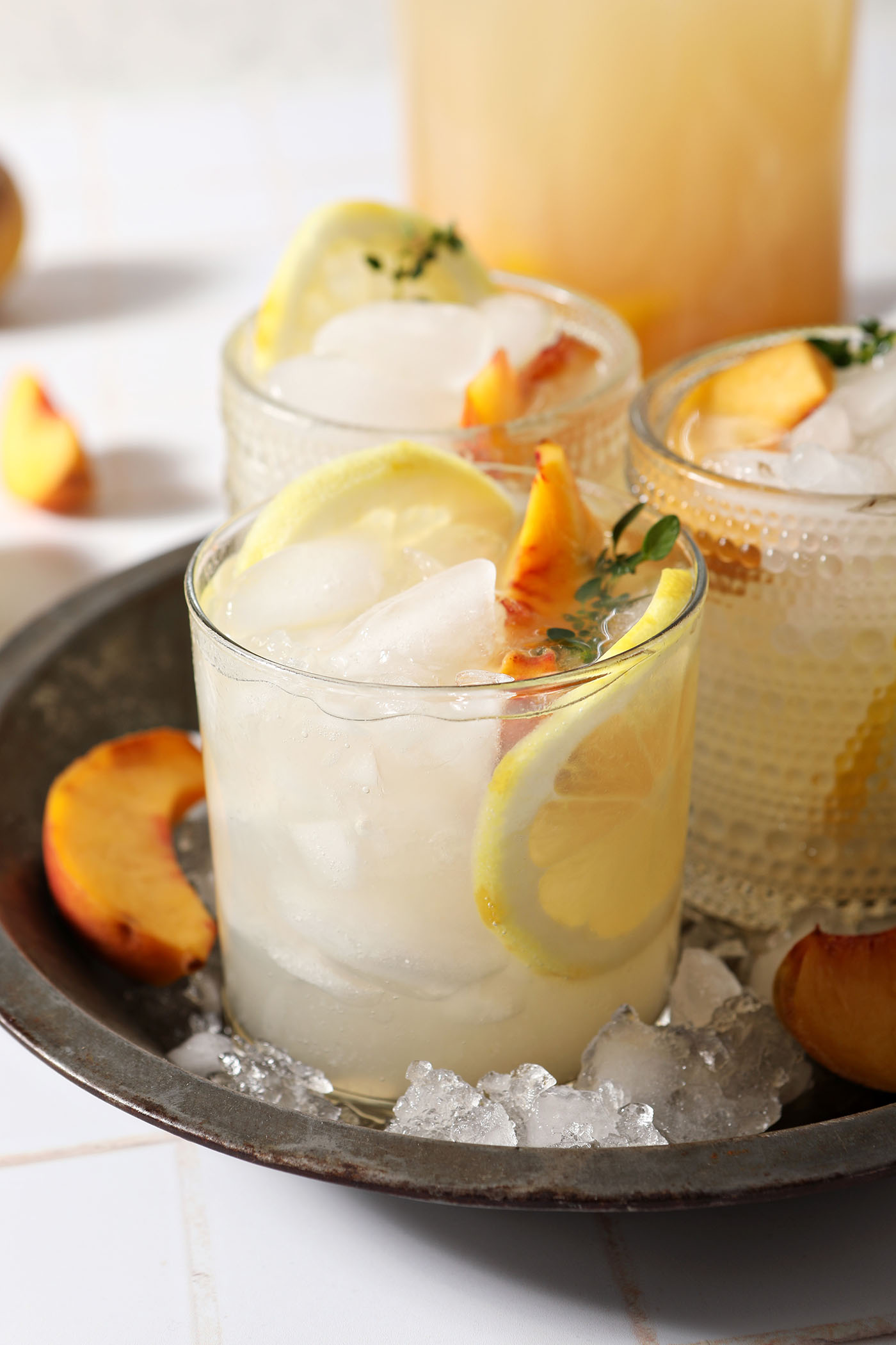 Three glasses of peach lemonade in a metal bowl in front of a pitcher of lemonade on a white tiled surface