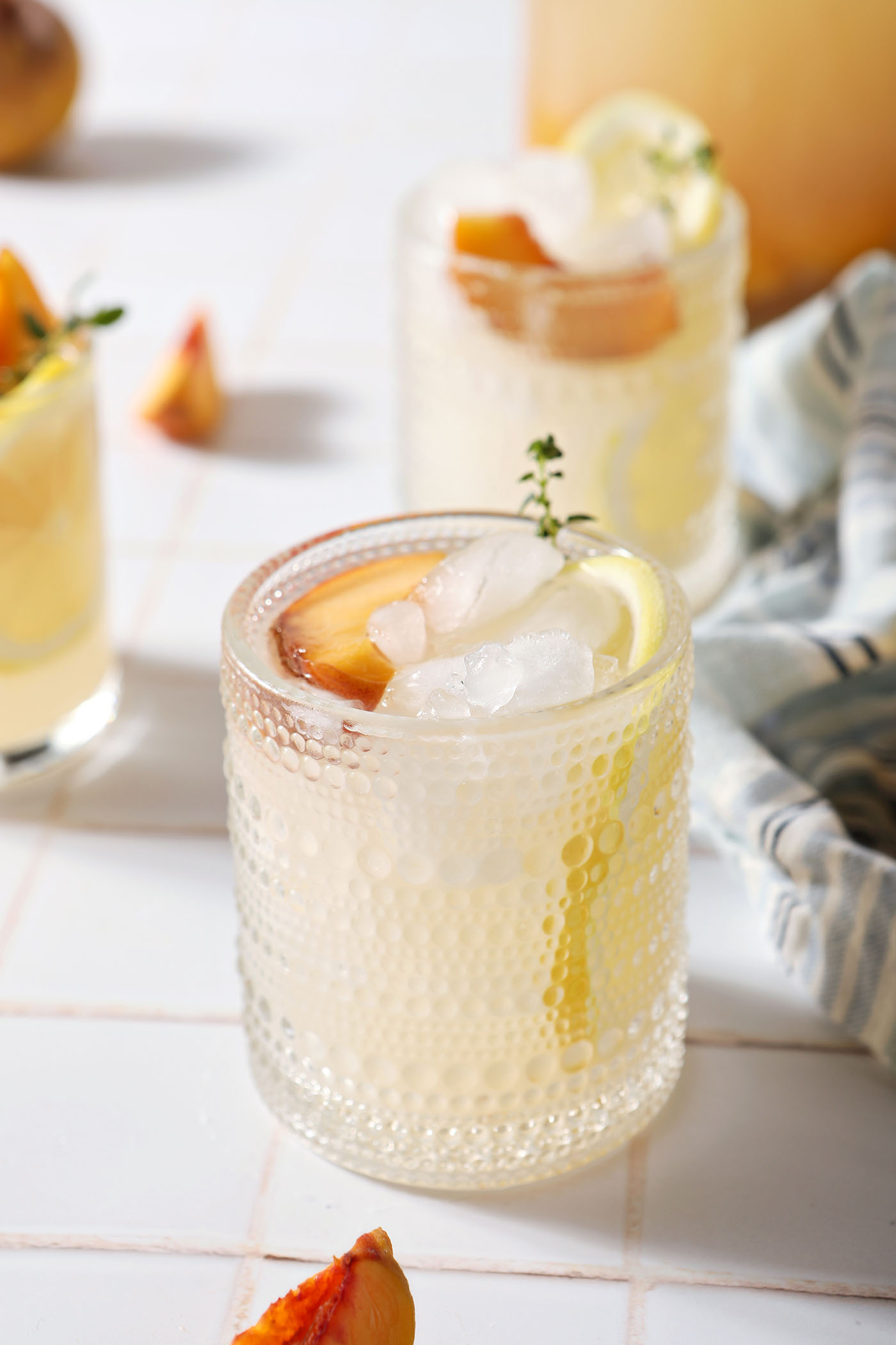 Three glasses of peach lemonade on a white tiled surface next to a blue striped linen in front of a pitcher and additional peaches