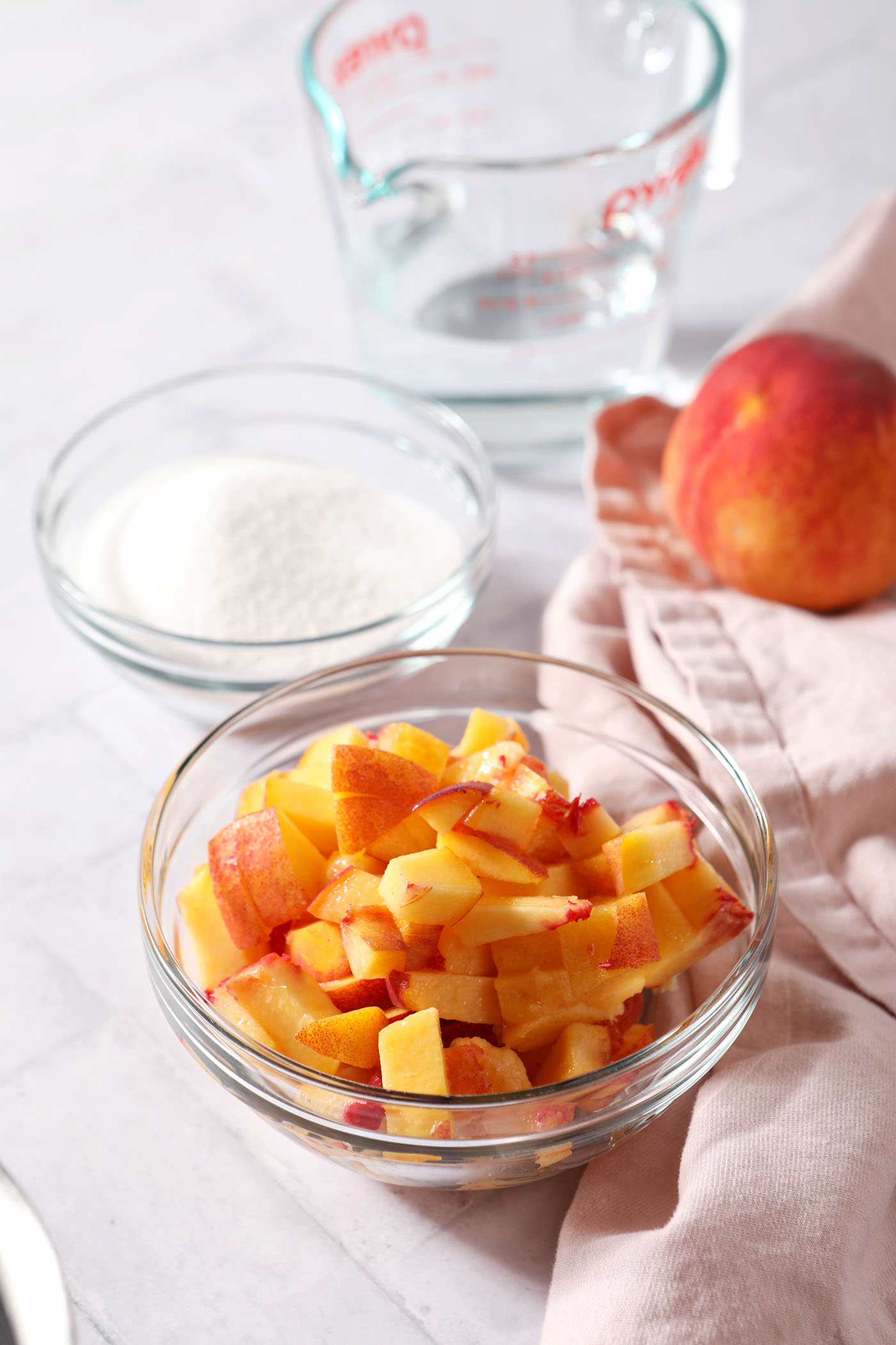 Ingredients to make peach syrup with fresh peaches in bowls on a grey tiled surface next to a pink linen and a fresh peach