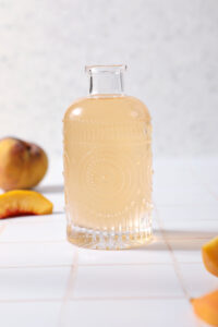 A bottle of peach simple syrup on a tiled surface in front of peaches