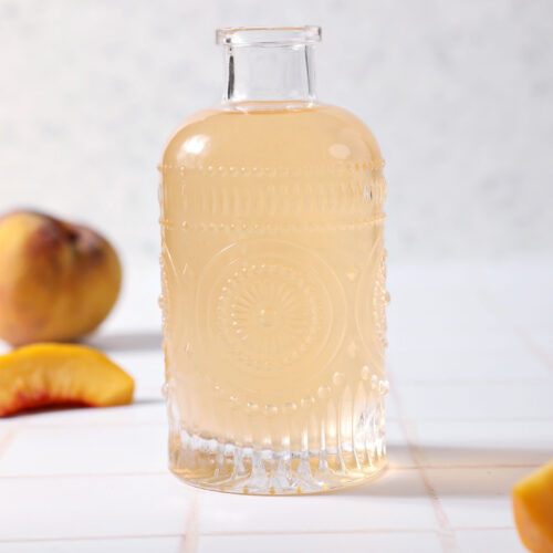 A bottle of peach simple syrup on a tiled surface in front of peaches