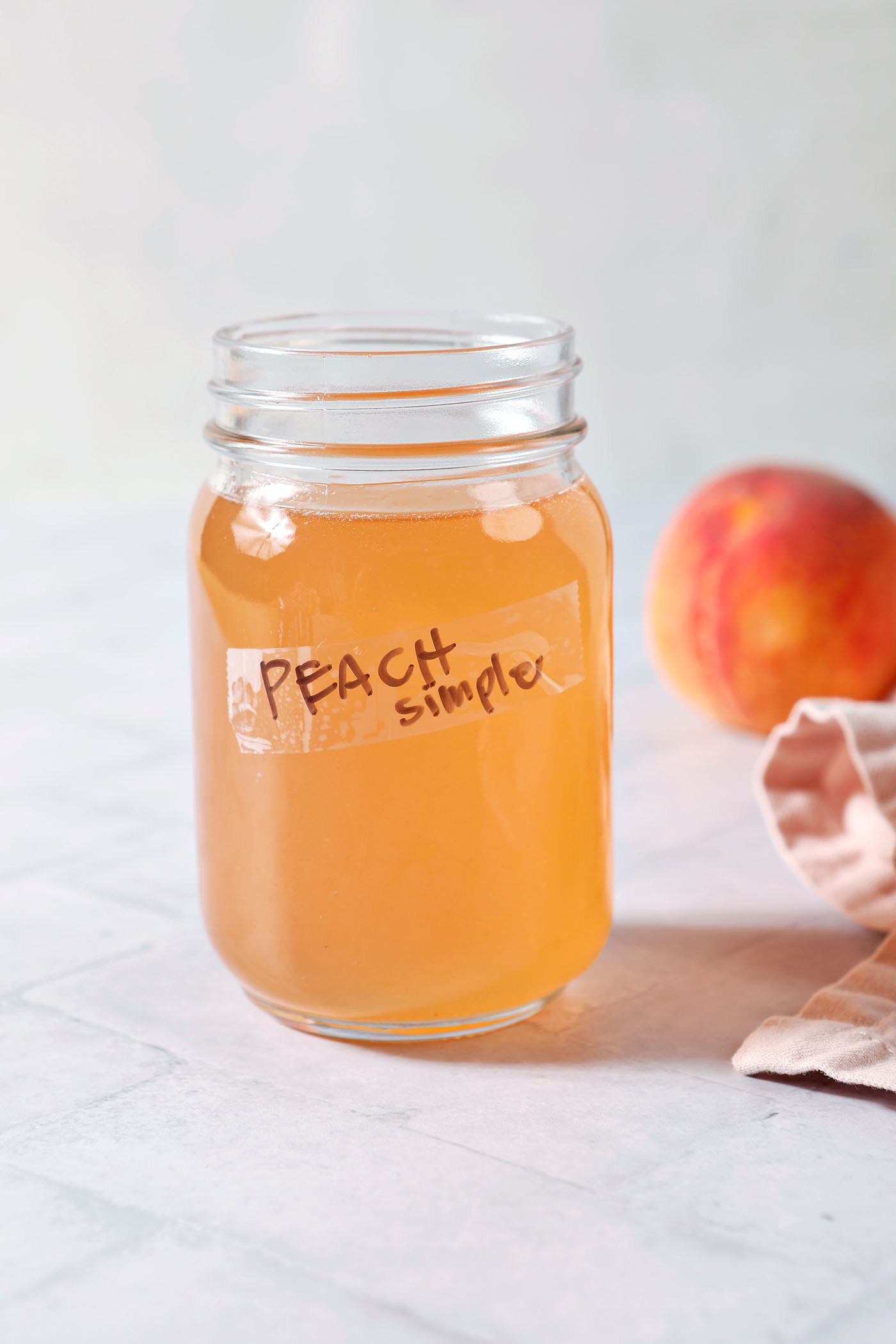 A jar of peach syrup labeled peach simple in front of a pink linen and a fresh peach on a grey tiled surface