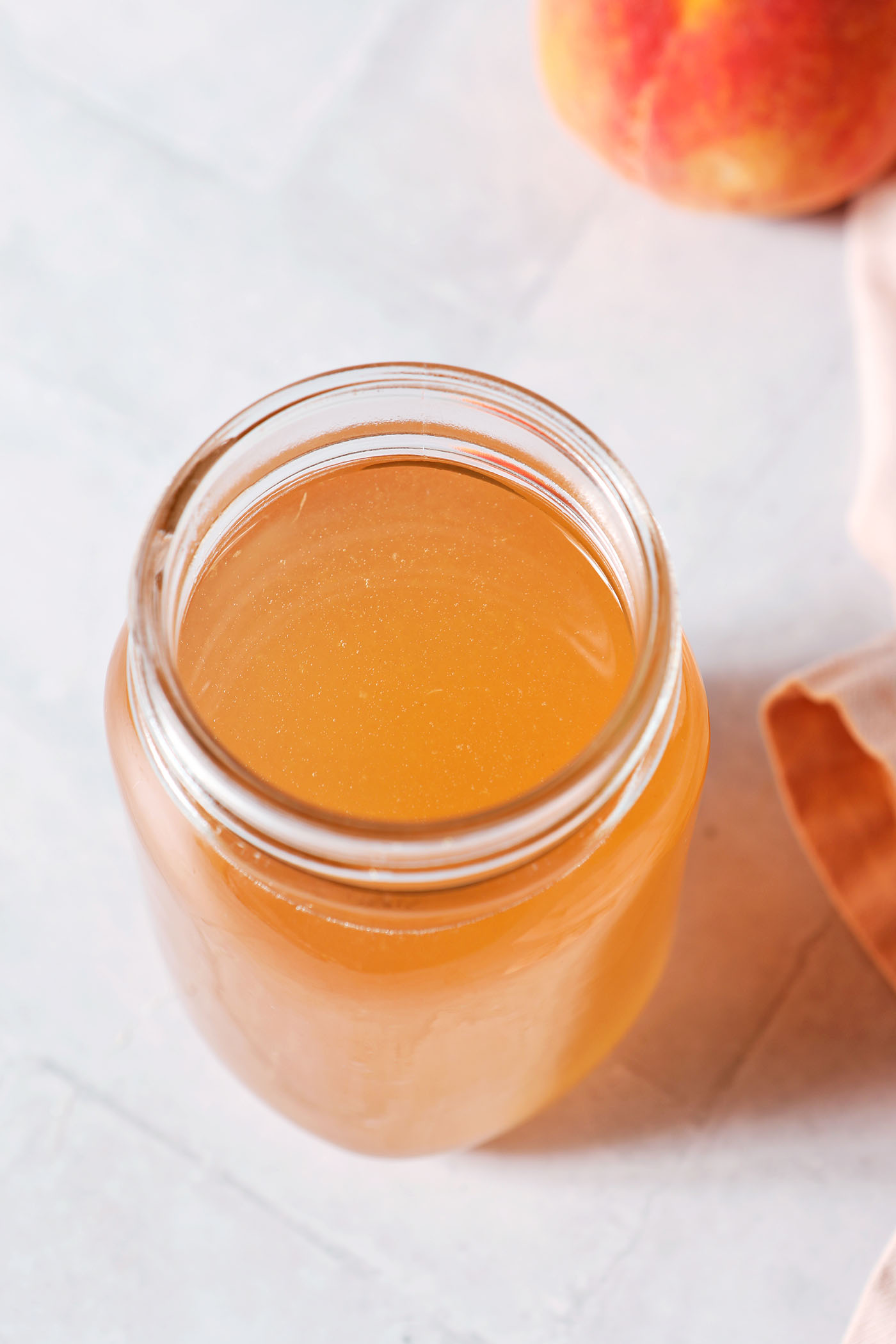 Overhead of a jar of peach simple syrup next to a peach and a pink linen on a grey tiled surface