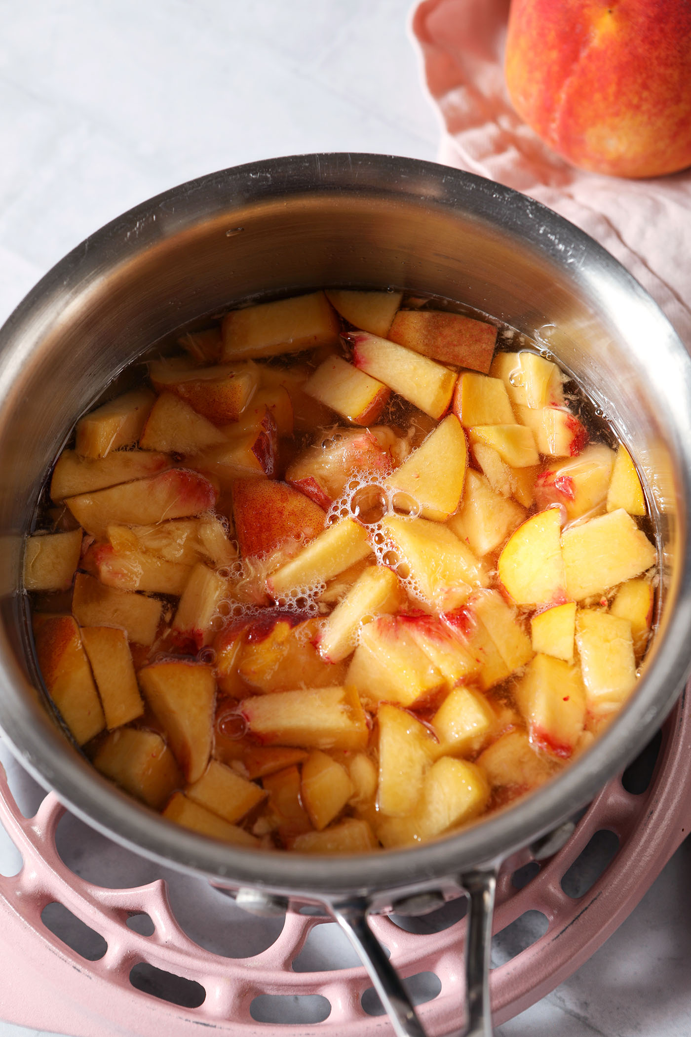 A saucepan of peaches in simple syrup on a pink trivet on a grey tiled surface