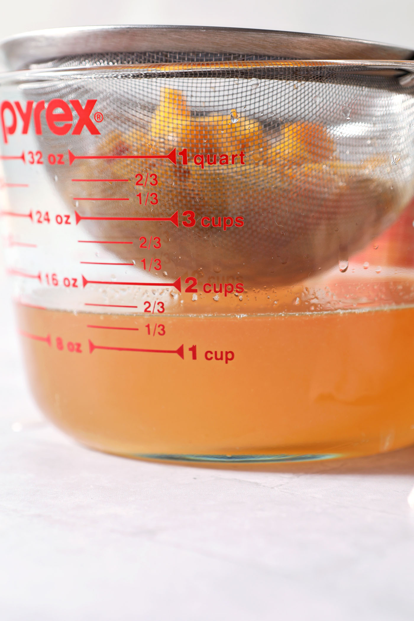 Syrup strains from peaches into a glass measuring cup on a grey tiled surface