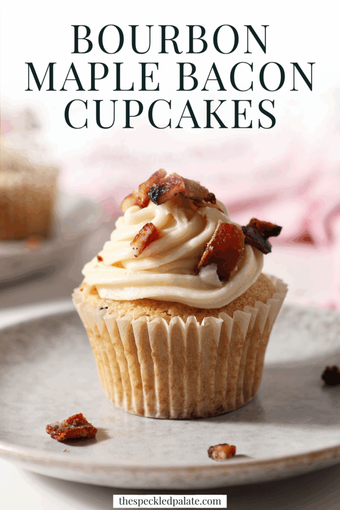 A cupcake topped with salted maple frosting and chopped maple bacon sits on a grey plate in front of a pink linen with the text bourbon maple bacon cupackes